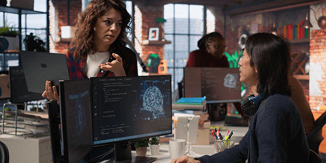 Two coworkers discuss code at a tech office with multiple monitors displaying programming tasks and a brain image on one screen.